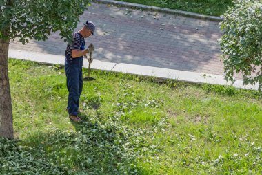 Yekaterinburg, Sverdlovsk Rusya - 07 25 2018: Çizgili mavi ve beyaz tişört ve mavi bir iş orta yaşlı erkeklerde pantolon ve kap mavi ve beyaz eldiven yan yatan yeşil yaprakları parkta bir komisyon ile yaz aylarında
