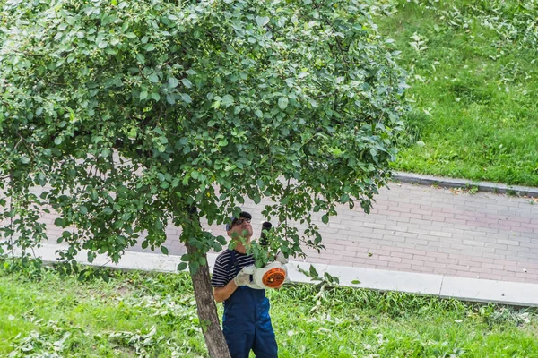 Yekaterinburg, Sverdlovsk Rusya - 07 25 2018: Çizgili mavi ve beyaz tişört ve mavi bir iş orta yaşlı erkeklerde pantolon ve kap ve beyaz eldiven inşaat parkta bir turuncu yaprak üfleyici ile yaz aylarında mavi