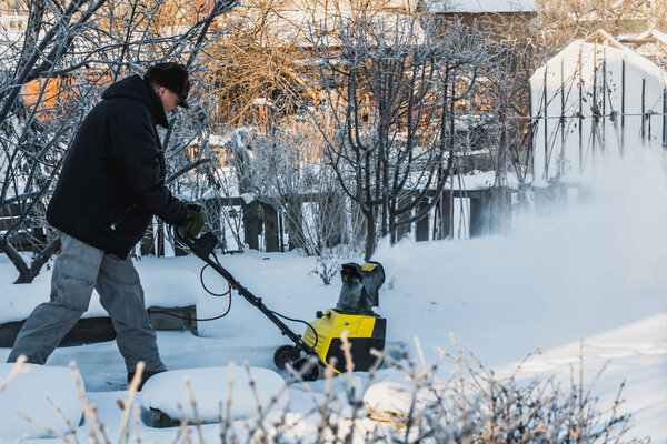 A man in a black jacket and a gray pants is brushing white snow with the yellow electric snow thrower in a winter garden