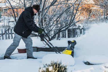 A man in a black jacket and a gray pants is brushing white snow with the yellow electric snow thrower in a winter garden clipart