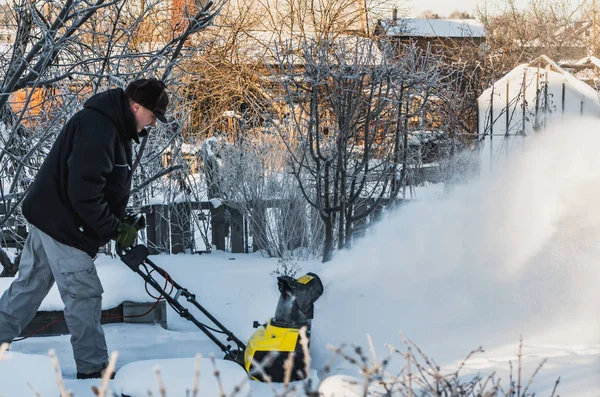 A man in a black jacket and a gray pants is brushing white snow with the yellow electric snow thrower in a winter garden