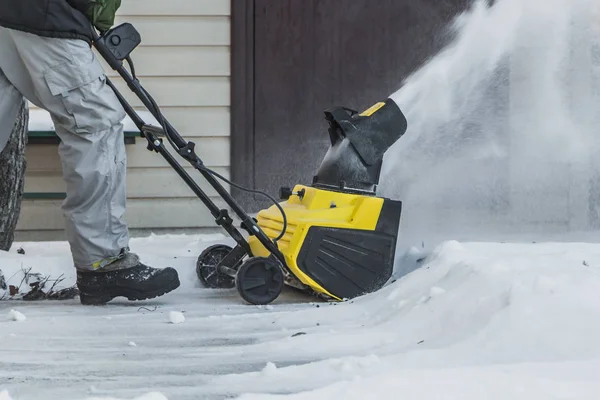 A man in a black jacket and a gray pants is brushing white snow with the yellow electric snow thrower in a winter garden