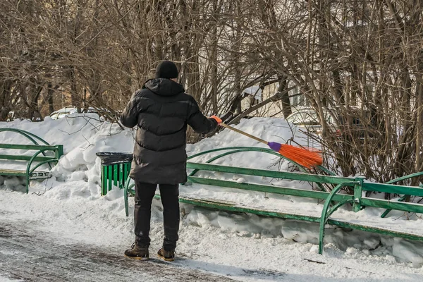 A man in a black clothes and orange gloves with a plastic bright orange broom in his hands sweeps the green bench from white snow on the street in winter