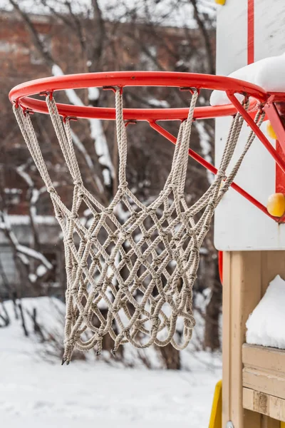 冬の庭の子供の遊び場に白いロープのネットと白い雪の美しい赤いバスケットボールのフープ。縦の写真 — ストック写真