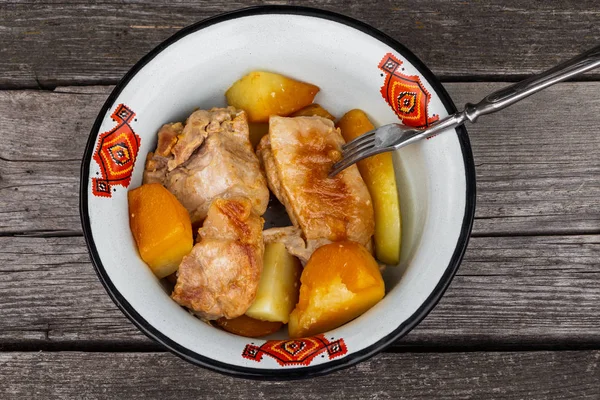 Juicy roast meat with potatoes served in a white metal bowl with a red ornament with a fork on an old rough gray wooden background