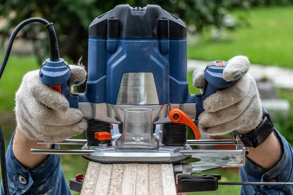 Man in werkende handschoenen snijdt een multiplex plaatrand met behulp van een blauwe elektrische frees tool met oranje hefboom en gele chips op een werkbank — Stockfoto