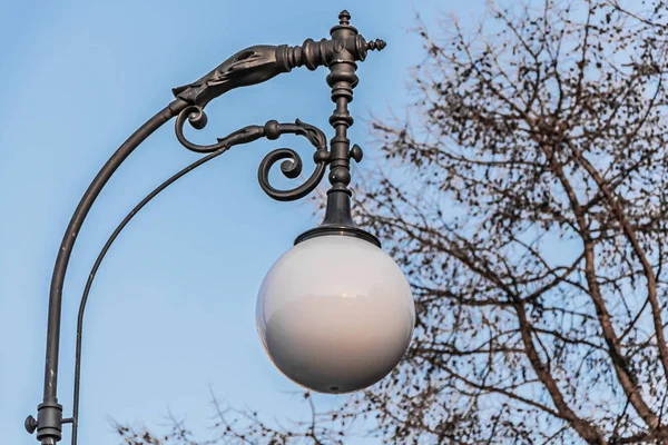 Una hermosa calle blanca esfera redonda linterna de iluminación con reflejos en un soporte de acero negro entre los árboles sin hojas en el fondo del cielo azul en un parque en otoño — Foto de Stock