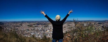 Dağın tepesinde kadının olduğu panoramik bir fotoğraf. Kolları açık ve şehre bakıyor. Minas Gerais 'deki Pocos de Caldas şehriyle dağda açık kollu bir kadının fotoğrafı.