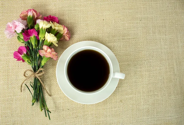 top view of black coffee in white cup with carnation bouquet tied with string bow on burlap