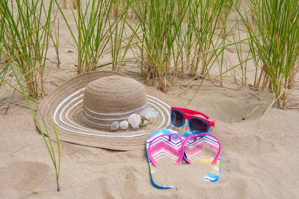 サングラスやフリップフロップでビーチの砂の貝殻で飾られた女性のスタイリッシュな夏の帽子 — ストック写真