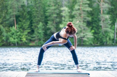 Yoga yapan genç bir Yogi kızı güzel bir gölün iskelesinde Tanrıça 'nın varyasyonlarıyla kıvrılıyor. Sağlıklı yaşam ve doğal denge kavramı 