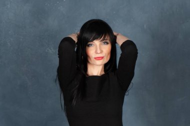 Etkileyici ve büyüleyici güzel bir kız, siyah elbiseli esmer. Güzel ve ince bir kadın kameraya bakıyor, gülümsüyor, tek renkli mavi, modaya uygun, pejmürde bir arka planda stüdyo portresi.