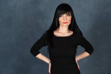 Etkileyici ve büyüleyici güzel bir kız, siyah elbiseli esmer. Güzel ve ince bir kadın kameraya bakıyor, gülümsüyor, tek renkli mavi, modaya uygun, pejmürde bir arka planda stüdyo portresi.