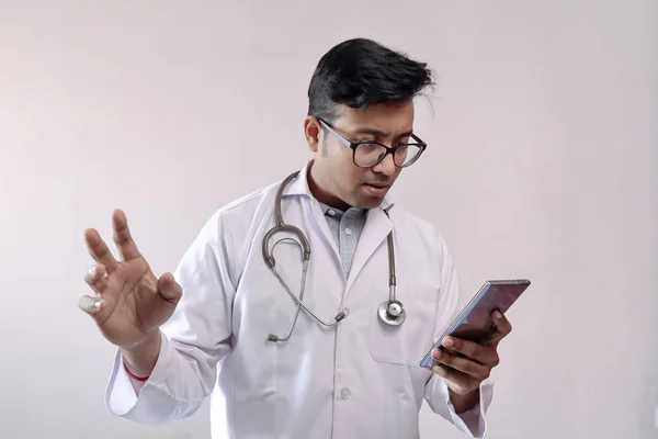 Male indian doctor in white coat and stethoscope in worried expression while looking at smart phone — Stock Photo, Image