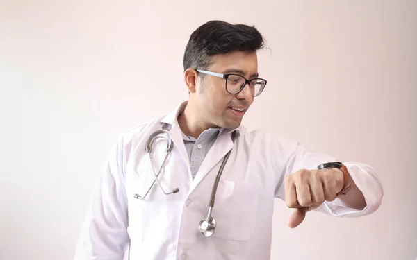 Αρσενικό ινδικό γιατρό με λευκό παλτό και στηθοσκόπιο βλέποντας έξυπνο ρολόι — Φωτογραφία Αρχείου