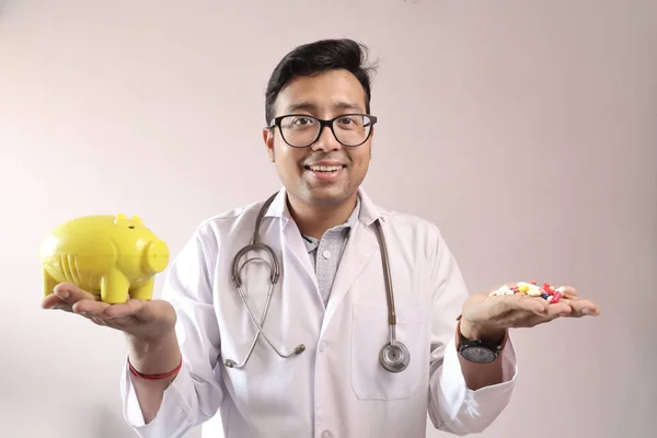 Médico indiano macho em casaco branco e estetoscópio com um banco piggy amarelo e pílulas de medicina na mão. imagem conceito despesa médica — Fotografia de Stock