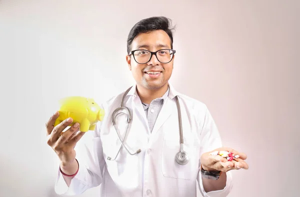 Médico indiano macho em casaco branco e estetoscópio com um banco piggy amarelo e pílulas de medicina na mão. imagem conceito despesa médica — Fotografia de Stock