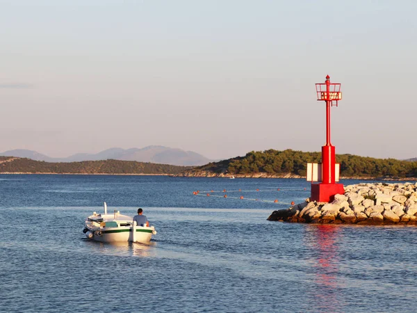 The fisherman on a boat swims out to catch fish in the rays of the early sun. A man on a boat sails past a stone breakwater with a red lighthouse and buoys to limit the beach area for bathing.