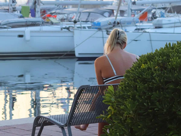 一位年轻女子坐在海滨公园的长凳上 在峡湾码头看游艇 一个穿着泳装的金发女郎在地中海港口有一个冥想 — 图库照片