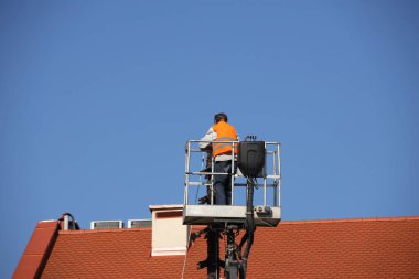 Bina mekanik kaldırma sepet içinde yükseklikte tulum işçi çalışıyor. Onarım ve inşaat kırmızı kiremit çatı üzerinde çalışıyorum. Yenileme tarihi bölümünde mimari eserlerinden.