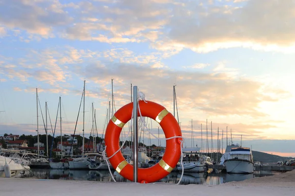 Orange life ring on the pier in the Croatian marina against the backdrop of sailing yachts. Safety on the water and saving drowning. Equipment of the seaport. Outfit of the rescue team