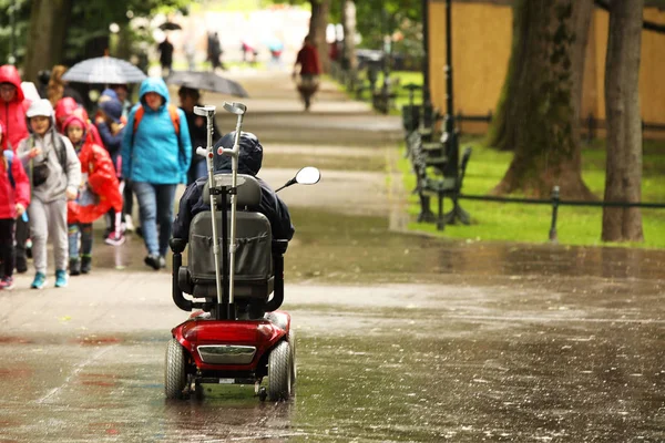 Ein älterer Herr im mechanischen Rollstuhl geht am Park vorbei an einer vorbeifahrenden Gruppe von Kindern. Mitgefühl und Hilfe für Menschen in Not. Aktive und Sozialisierung von Menschen mit Behinderungen — Stockfoto