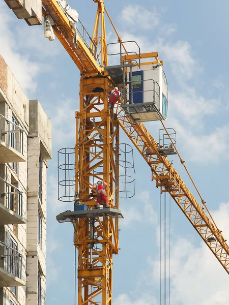Trabalhadores guindastes em macacões estão reparando um guindaste de torre de arranha-céus trabalhando durante a construção de um edifício de vários andares. Trabalho arriscado em altura. Levantamento de materiais de construção pesados. Desenvolvimento da cidade. — Fotografia de Stock