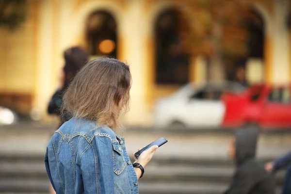 A young girl in a denim jacket is sitting on talking on a mobile phone. Modern communications and electronic communications technology. Communication between people. City street under the sun