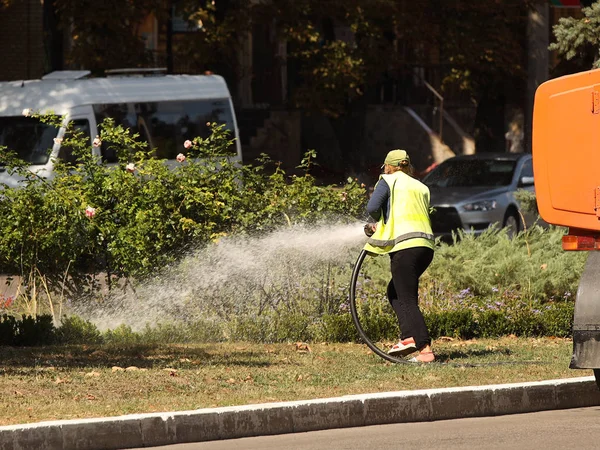Femeie oraș îmbunătățire lucrător stropește apă pe un gazon verde cu flori într-o zi însorită fierbinte. Lupta împotriva secetei și salvarea spațiilor verzi. Grija pentru peisajul decorativ. Transport de apă — Fotografie, imagine de stoc