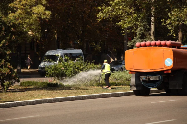 Una mujer trabajadora de mejoras en la ciudad rocía agua en un césped verde con flores en un día soleado y caluroso. Lucha contra la sequía y rescate de espacios verdes. Cuidado del paisaje decorativo. Transporte por agua — Foto de Stock