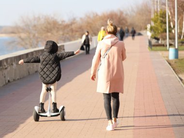 Genç bir anne ve kız gezinti güvertesinde yürüyor. Jiroskopla parkta gezen bir kız. Modern elektrik ekolojik taşıma yöntemi. Şehirdeki ekoloji. Çocukların etkinliği. Sağlık sporları.