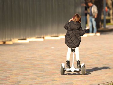 Parkta bir kız jiroskopla telefonda konuşuyor. Modern elektro-ekolojik taşıma yöntemi. Şehirdeki ekoloji. Çocukların etkinliği. Sağlık için spor. Dengeni koruyabilmen için.