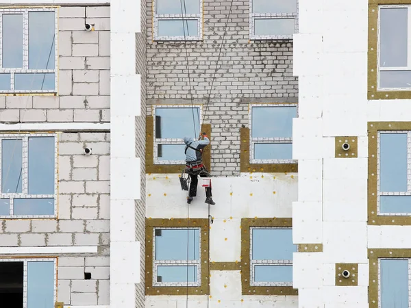 A builder in climbing equipment insulates the facade of a high-rise building with foam plastic. Safety measures for high-altitude construction work. Heat conservation in homes and energy savings.