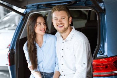 Onların satın hakkında mutlu. Güzel genç sevgi dolu kadın yakışıklı mutlu kocası smiling birlikte yeni bir arabanın bagajında otururken kameraya gülüyor