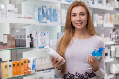 Happy beautiful woman smiling to the camera joyfully, holding products she is buying at the drugstore. Cheerful female customer posing at the pharmacy, holding two product bottles, copy space