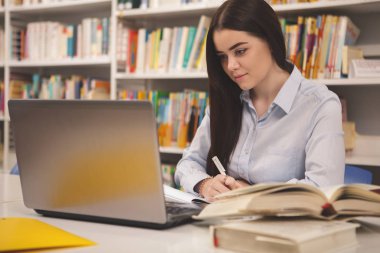 Beautiful female student taking notes in her textbook, sitting in front of the laptop, copy space. Attractivr woman studying at college library, using her computer. Online research, homework project concept