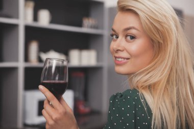 Mutfak evde şarap içme onun omuz üzerinden kameraya gülümseyen bir muhteşem mutlu sarışın saçlı kadının kapatın. Güzel kadın bir kadeh şarap ile kutluyor. Yaşam tarzı, gayrimenkul konsept