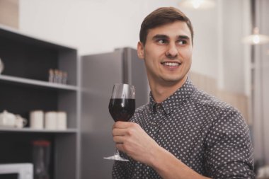 Neşeyle gülümseyerek, uzakta, kopya alanı arıyor mutfakta şarap içme, akşamları evde rahatlatıcı mutlu çekici bir adam. kırmızı şarap tadımı, modern evinde istirahat yakışıklı adam
