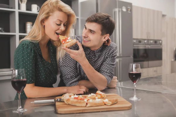 Handsome cheerful man offering a bite of pizza to his gorgeous girlfriend. Beautiful happy woman biting piece of pizza, her husband is holding. Couple having dinner in the kitchen, copy space. Love concept