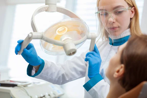 Diş onun hastanın incelemek hazırlanıyor diş lamba, ayarlama çekici kadın diş hekimi. Onun Kliniği'nde çalışan profesyonel diş hekimi. Kadın ziyaret dişçi diş muayenesi için. Meslek kavramı