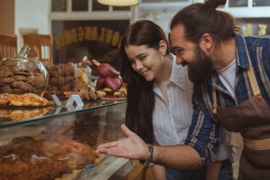 Young beautiful woman buying delciious freshly baked pastry at local bakery. Professional baker pointing at the display, talking to his customer. Bearded mature man working at his cafe, selling desserts clipart