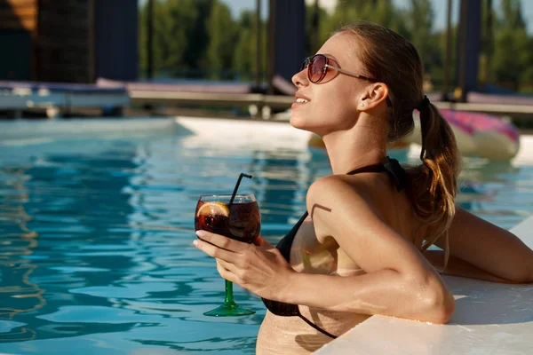 Stunning beautiful young woman enjoyoing sunbathing in the pool with a drink in her hand, copy space. Cheerful attractive woman having  cocktail at the poolside. Relation, lifestyle, summertime concept