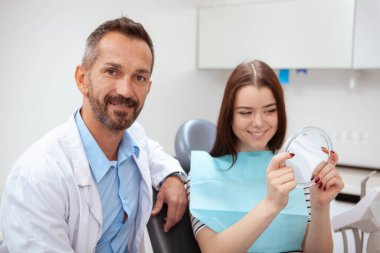 Kameraya gülümseyen yakışıklı erkek doktor, aynaya gülümseyen kadın hasta. Profesyonel diş tedavisinden sonra dişlerini aynada kontrol eden genç bir kadın.