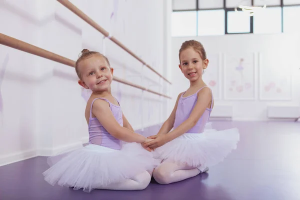 Lovely little ballerinas holding hands, sitting on the floor at ballet school, smiling to the camera. Happy adorable young ballet dancing girls resting after exercising