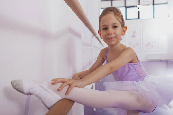 Charming young ballerina smiling to the camera, stretching on ballet barre at dance school, copy space