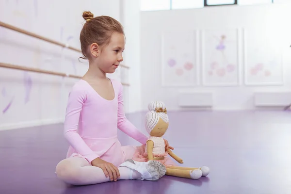 Charming young ballerina in pink leotard sitting on the floor with her ballerina doll, resting at ballet school after exercising