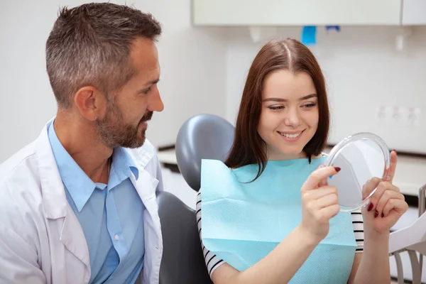 Happy beautiful woman examining her healthy white teeth in the mirror. Attractive female patient looking in the mirror after dental treatment by professional dentist