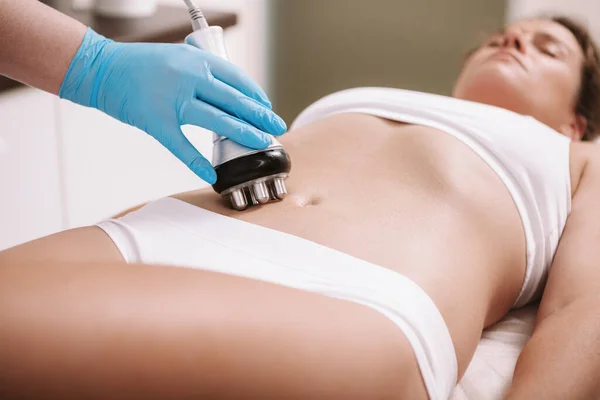Güzellik kliniğinde karın derisi üzerinde rf kaldırma işleminden hoşlanan bir kadın. Profesyonel güzellik uzmanı. Rf-lift makinesi kullanıyor. Kadın bir müşterinin karnını sıkıyor.