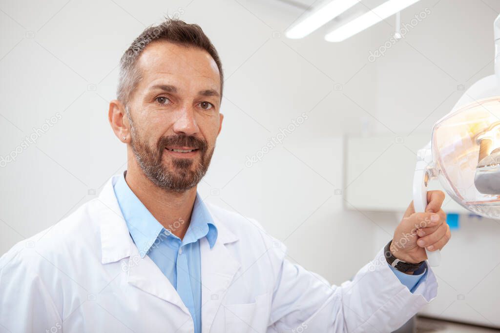 Handsome mature male dentist smiling to the camera, holding dental lamp, copy space