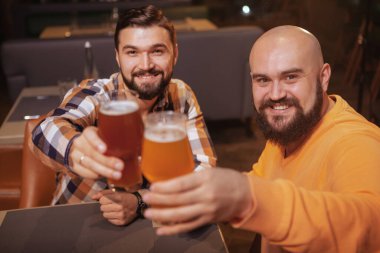 Two cheerful handsome men smiling to the camera, clinking their beer glasses clipart